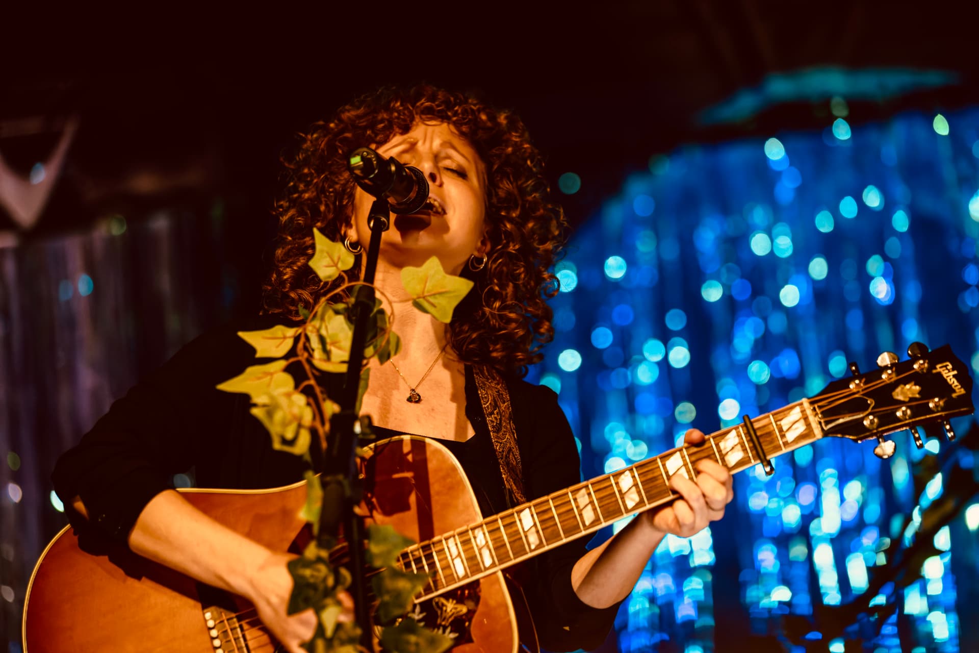 Stina Bekker i nærbillede under koncert Nærbillede af Stina Bekker, der synger med guitar foran blå bokeh-lys til koncert